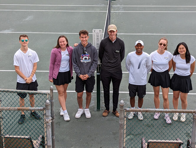 Seven people standing shoulder to shoulder at a tennis court looking at camera and smiling